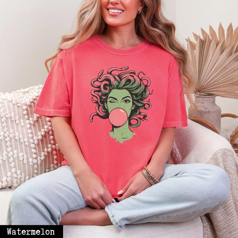 May include: A watermelon-coloured t-shirt with a graphic of Medusa winking and blowing a bubblegum bubble. The Medusa illustration has green hair and a green face. The word "Watermelon" is printed at the bottom of the image.