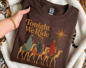 Könnte beinhalten: Ein dunkelbraunes Sweatshirt mit Rundhalsausschnitt und dem Schriftzug Tonight We Ride in goldener Schrift. Die Grafik zeigt drei Figuren mit Kronen und Gewändern, die jeweils auf einem Kamel reiten, unter einem großen goldenen Stern.