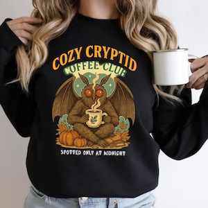 May include: Black sweatshirt featuring the text "COZY CRYPTID COFFEE CLUB" and a cryptid illustration holding a coffee cup. The design includes pumpkins, leaves, and the phrase "SPOTTED ONLY AT MIDNIGHT".