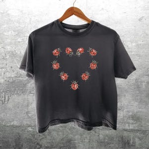 May include: A charcoal gray short-sleeve t-shirt featuring a graphic print of seven red ladybugs with black spots arranged in the shape of a heart on the center of the chest. The shirt has a crew neckline and a standard fit with hemmed sleeves.