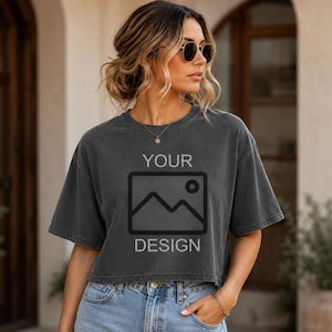 May include: A charcoal gray cropped t-shirt featuring a black graphic of a mountain landscape inside a rectangular frame with the text YOUR above and DESIGN below the graphic. The shirt has short sleeves and a crew neckline.