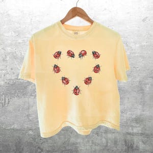 May include: A pale yellow short-sleeve t-shirt featuring a graphic print of red and black ladybugs arranged in the shape of a heart on the center of the chest. The shirt has a crew neckline and is displayed on a wooden hanger against a gray background.
