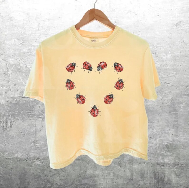 May include: A pale yellow short-sleeve t-shirt featuring a graphic print of red and black ladybugs arranged in the shape of a heart on the center of the chest. The shirt has a crew neckline and is displayed on a wooden hanger against a gray background.