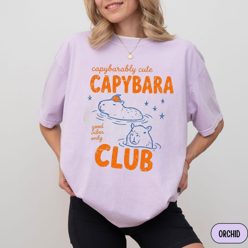 May include: A light purple t-shirt featuring a graphic print with the text "capybarably cute CAPYBARA CLUB" and "good vibes only". The design includes two blue line-art capybaras in water, one with an orange fruit on its head, and blue stars.