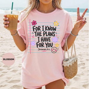 May include: A light pink t-shirt featuring blue horizontal lines and the black text: FOR I KNOW THE PLANS I HAVE FOR YOU Jeremiah 29:11. The design includes colorful flower and star illustrations, along with a small yellow smiley face icon.