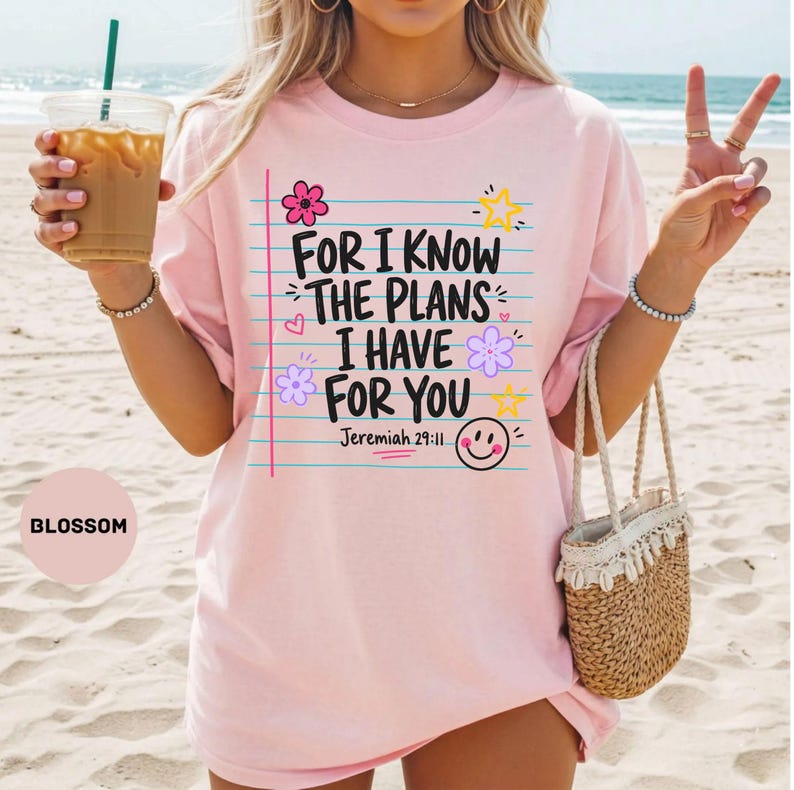 May include: A light pink t-shirt featuring blue horizontal lines and the black text: FOR I KNOW THE PLANS I HAVE FOR YOU Jeremiah 29:11. The design includes colorful flower and star illustrations, along with a small yellow smiley face icon.