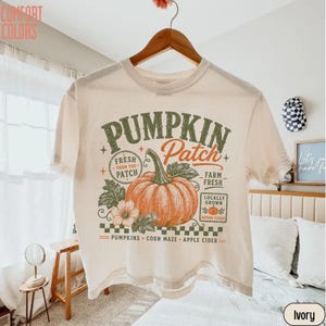 May include: An ivory short-sleeve t-shirt featuring a graphic print of an orange pumpkin with green leaves. The text reads PUMPKIN Patch, FRESH FROM THE PATCH, FARM FRESH, LOCALLY GROWN, PUMPKINS, CORN MAZE, and APPLE CIDER in green and orange.