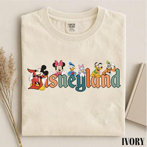 May include: Ivory-colored t-shirt featuring the word "Disneyland" in colorful retro lettering, with cartoon characters Mickey Mouse, Minnie Mouse, Donald Duck, Daisy Duck, Pluto, and Goofy. The shirt is folded and has a tag.