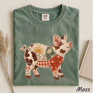 May include: A moss green t-shirt featuring a graphic of a pig composed of various patchwork patterns including a sunburst, daisies, brown cow spots, red gingham, and floral prints. The inner neck label displays the text Comfort Colors.