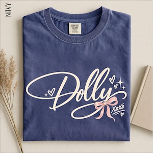 May include: A navy blue crew neck t-shirt featuring the word Dolly in a large, cream-colored cursive script. The design includes a light pink bow, small heart outlines, and star accents, with the text XOXO written in a smaller script below the bow.