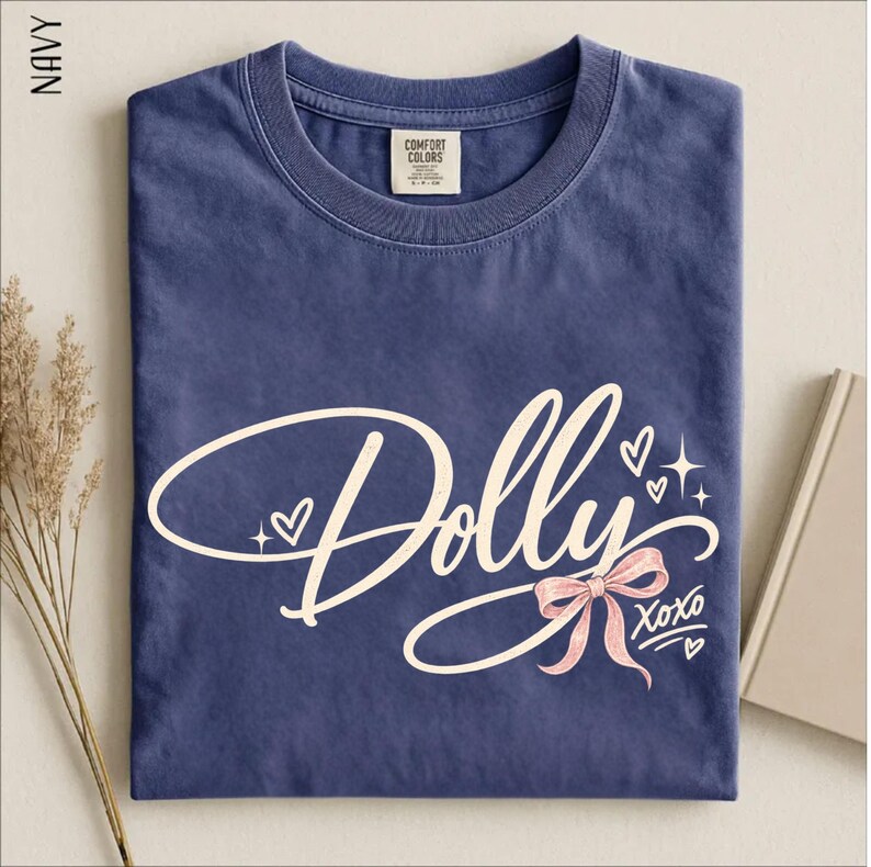May include: A navy blue crew neck t-shirt featuring the word Dolly in a large, cream-colored cursive script. The design includes a light pink bow, small heart outlines, and star accents, with the text XOXO written in a smaller script below the bow.
