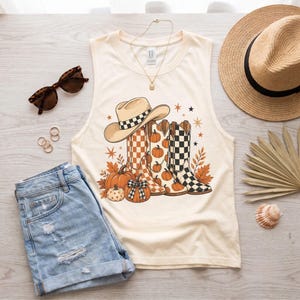 May include: A cream-colored sleeveless tank top featuring a graphic print of three cowboy boots, pumpkins, and a cowboy hat. The boots display checkered and pumpkin patterns. The design includes orange autumn leaves and small star shapes.