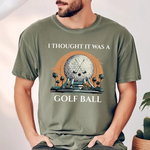 Puede incluir: Camiseta verde oliva con un gráfico de una pelota de golf, palos de golf cruzados y el texto "I THOUGHT IT WAS A GOLF BALL". El gráfico de la pelota de golf está sobre un fondo naranja.