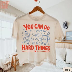 May include: An ivory short-sleeve t-shirt featuring a graphic print with the red text "YOU CAN DO" and "HARD THINGS" surrounding blue cartoon illustrations of a pencil, an apple, and a book, with the blue text "you got this!" in the center.