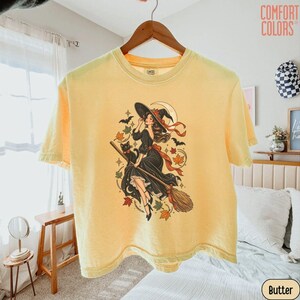 May include: A pale yellow short-sleeve t-shirt featuring a graphic print of a person in a black dress and witch hat riding a broomstick with a black cat, bats, stars, and autumn leaves. The text COMFORT COLORS is printed in the top right corner.