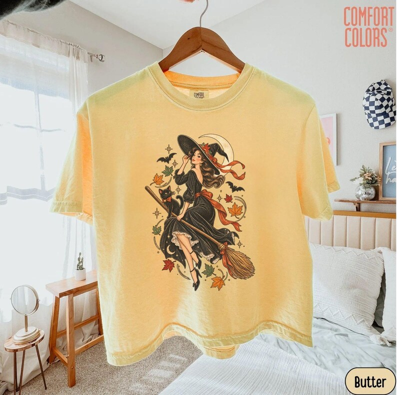 May include: A pale yellow short-sleeve t-shirt featuring a graphic print of a person in a black dress and witch hat riding a broomstick with a black cat, bats, stars, and autumn leaves. The text COMFORT COLORS is printed in the top right corner.