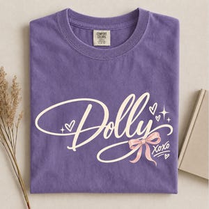 May include: A purple short-sleeved t-shirt featuring the word Dolly in a cream-colored cursive script. The design includes a small pink bow, several hearts, and star accents, with the text XOXO written in a matching cream-colored script.