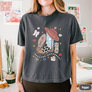 May include: A charcoal gray short-sleeve t-shirt featuring a graphic print of a cowboy boot with a cow print pattern, a brown hat, a pink bow, and blocks with the text "TAYLOR" and "13". The design includes small stars and floral accents.