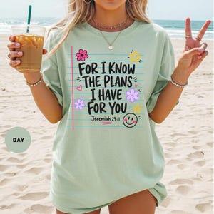 May include: A light green t-shirt featuring a graphic design that resembles a piece of lined notebook paper. The text reads, "FOR I KNOW THE PLANS I HAVE FOR YOU Jeremiah 29:11," accompanied by colorful illustrations of flowers, stars, and a smiley face.
