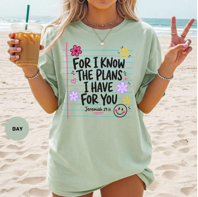 May include: A light green t-shirt featuring a graphic design that resembles a piece of lined notebook paper. The text reads, "FOR I KNOW THE PLANS I HAVE FOR YOU Jeremiah 29:11," accompanied by colorful illustrations of flowers, stars, and a smiley face.