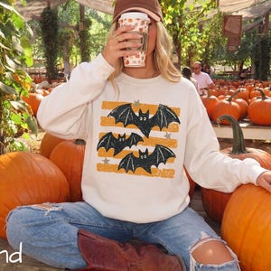 May include: A sand-colored crewneck sweatshirt featuring a graphic print of three black bats flying over horizontal orange distressed stripes and small gray stars. The long-sleeved pullover is worn with blue denim jeans and brown boots.