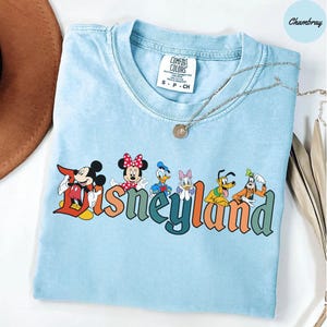 May include: Light blue Comfort Colors t-shirt featuring the word "Disneyland" in colorful letters, with Mickey Mouse, Minnie Mouse, Donald Duck, Daisy Duck, Goofy, and Pluto graphics. A silver necklace with a pendant is draped across the shirt.