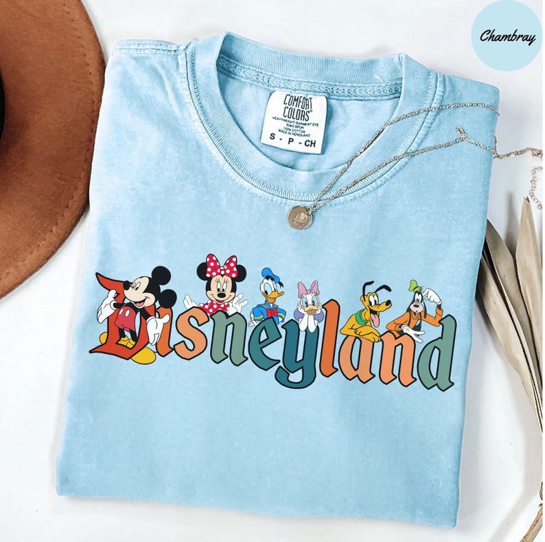 May include: Light blue Comfort Colors t-shirt featuring the word "Disneyland" in colorful letters, with Mickey Mouse, Minnie Mouse, Donald Duck, Daisy Duck, Goofy, and Pluto graphics. A silver necklace with a pendant is draped across the shirt.