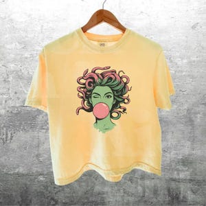 May include: Pale yellow t-shirt featuring a graphic of a woman with green skin, pink bubblegum, and snakes for hair. The t-shirt is hanging on a wooden hanger against a grey textured wall.