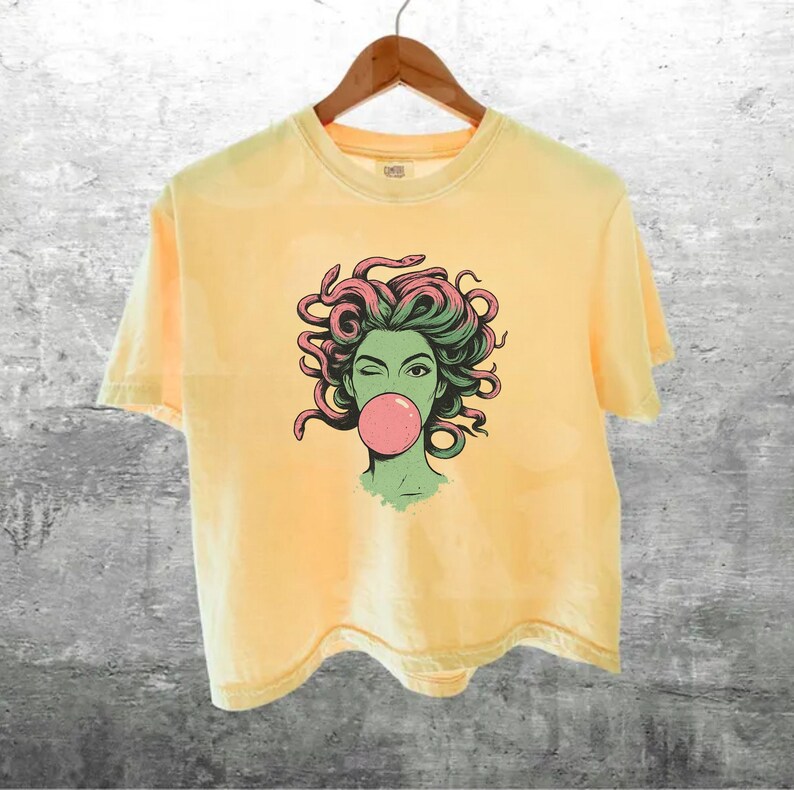 May include: Pale yellow t-shirt featuring a graphic of a woman with green skin, pink bubblegum, and snakes for hair. The t-shirt is hanging on a wooden hanger against a grey textured wall.