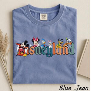 May include: A blue Comfort Colors t-shirt featuring the word "Disneyland" in colorful letters with Disney characters Mickey Mouse, Minnie Mouse, Donald Duck, Daisy Duck, Pluto, and Goofy. The shirt is labeled "Blue Jean".