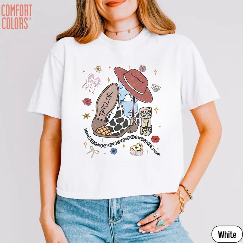 May include: A white short-sleeve t-shirt featuring a graphic print of a brown cowboy hat, a cow-print western boot with the name Taylor and number 13, stacked blocks with letters, a bow, flowers, stars, and a beaded string design on the front.