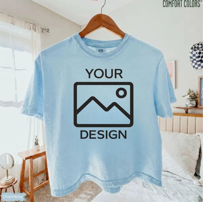 May include: A light blue short-sleeve t-shirt hanging on a wooden hanger. The front of the shirt features the black text "YOUR" above a black graphic icon of a mountain landscape inside a rectangle, with the black text "DESIGN" below it.