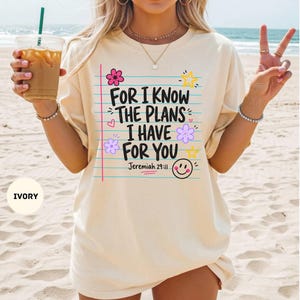 May include: An ivory short-sleeve t-shirt featuring a graphic print of blue horizontal lines resembling notebook paper. The text reads, "FOR I KNOW THE PLANS I HAVE FOR YOU Jeremiah 29:11," accompanied by pink and purple flowers, yellow stars, and a smiley face.