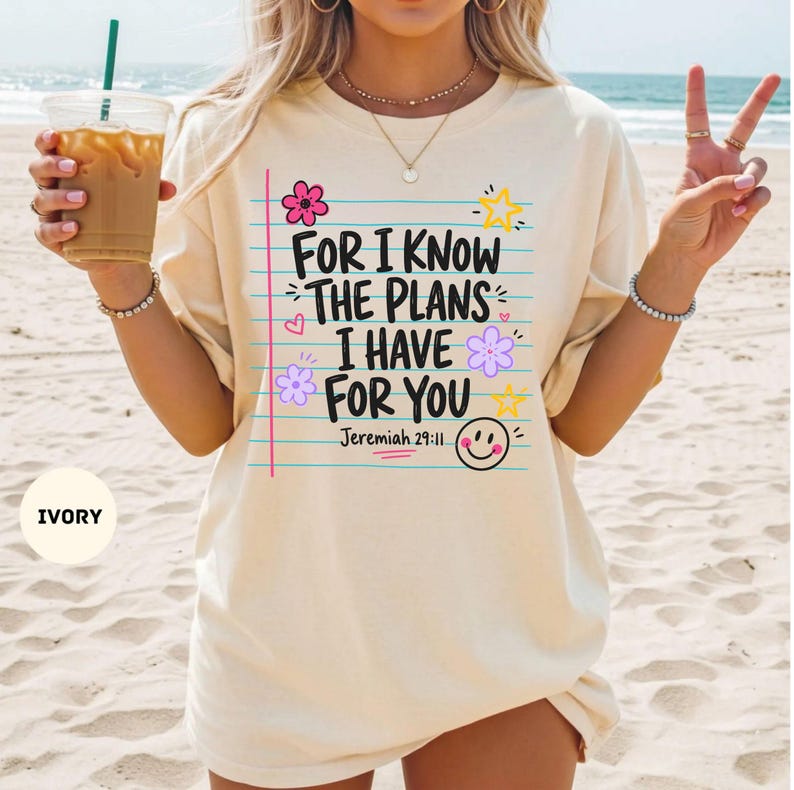May include: An ivory short-sleeve t-shirt featuring a graphic print of blue horizontal lines resembling notebook paper. The text reads, "FOR I KNOW THE PLANS I HAVE FOR YOU Jeremiah 29:11," accompanied by pink and purple flowers, yellow stars, and a smiley face.
