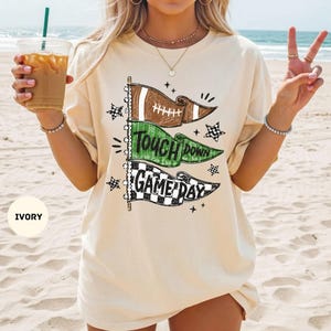 May include: An ivory short-sleeve t-shirt featuring a graphic print of three stacked pennant flags. The top flag is brown with a football design, the middle green flag reads "TOUCH DOWN", and the bottom black and white checkered flag reads "GAME DAY".