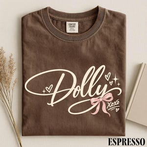 May include: A brown short-sleeve t-shirt with a crew neckline. The front features the word Dolly in large cream script, accented by small hearts, starbursts, a light pink ribbon bow, and the text XOXO. The inner neck label reads Comfort Colors.