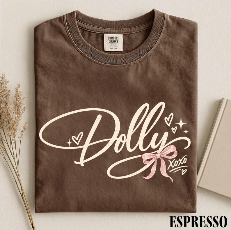 May include: A brown short-sleeve t-shirt with a crew neckline. The front features the word Dolly in large cream script, accented by small hearts, starbursts, a light pink ribbon bow, and the text XOXO. The inner neck label reads Comfort Colors.