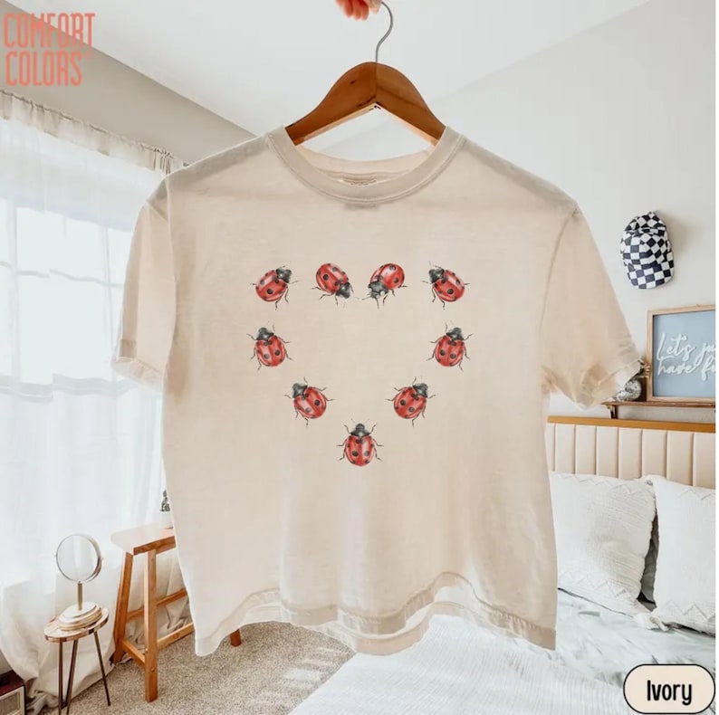 May include: An ivory short-sleeve t-shirt featuring a graphic print of red ladybugs arranged in the shape of a heart on the center chest. The shirt has a crew neckline and is displayed on a wooden hanger. The text COMFORT COLORS is visible.