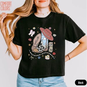 May include: A black short-sleeve t-shirt featuring a graphic print of a cowboy boot with a cow print pattern, a brown hat, a pink bow, and blocks with the number 13. The word TAYLOR is printed on the boot. The shirt has a Comfort Colors logo.