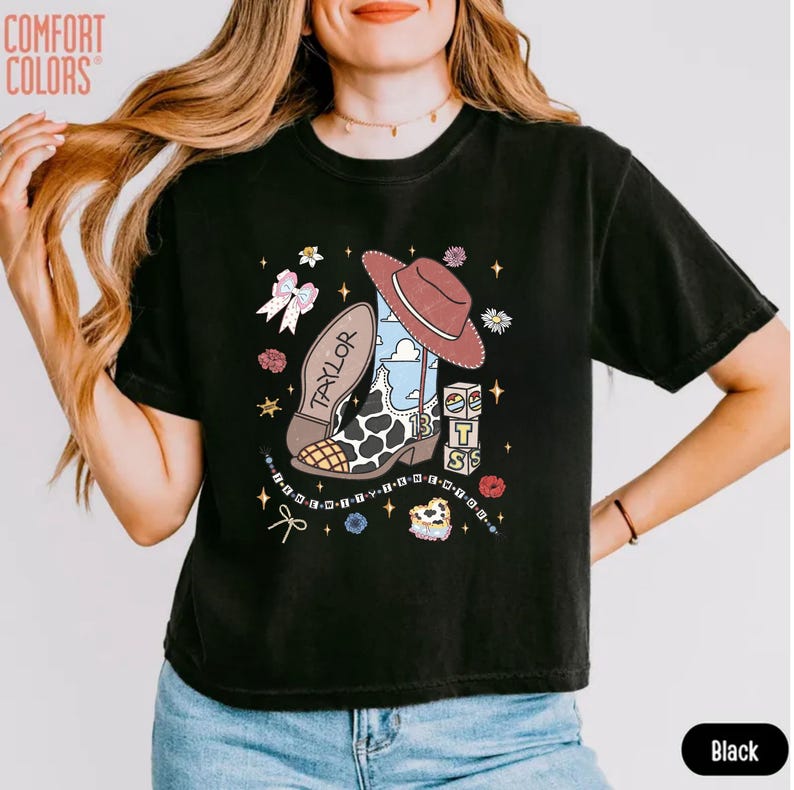 May include: A black short-sleeve t-shirt featuring a graphic print of a cowboy boot with a cow print pattern, a brown hat, a pink bow, and blocks with the number 13. The word TAYLOR is printed on the boot. The shirt has a Comfort Colors logo.