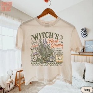 May include: An ivory short-sleeve t-shirt featuring a graphic print with the text "Witch's Herb Garden" and illustrations of herbs, mushrooms, a potion bottle, and a crescent moon. The design includes the words "Freshly Gathered," "Locally Grown Herbs," "Moonlit Grown," "Sage," "Potions," and "Moonlight."