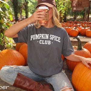 May include: A charcoal grey short-sleeve t-shirt featuring the white text PUMPKIN SPICE CLUB printed on the chest. The shirt is worn with light blue distressed denim jeans and brown leather boots, paired with a brown baseball cap.