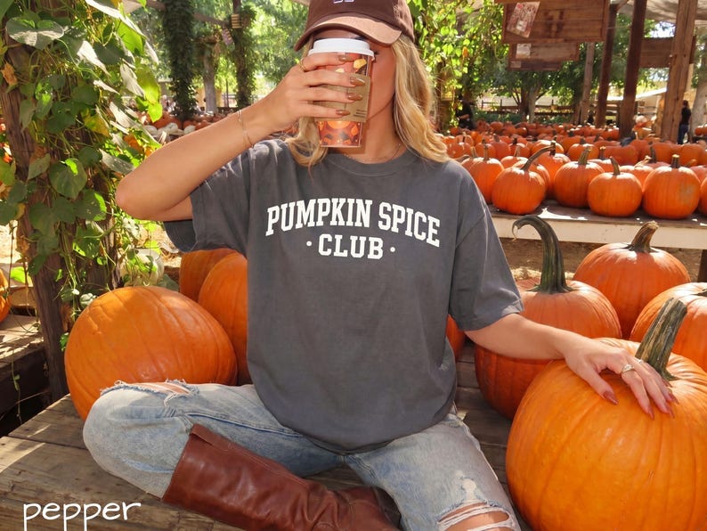 May include: A charcoal grey short-sleeve t-shirt featuring the white text PUMPKIN SPICE CLUB printed on the chest. The shirt is worn with light blue distressed denim jeans and brown leather boots, paired with a brown baseball cap.