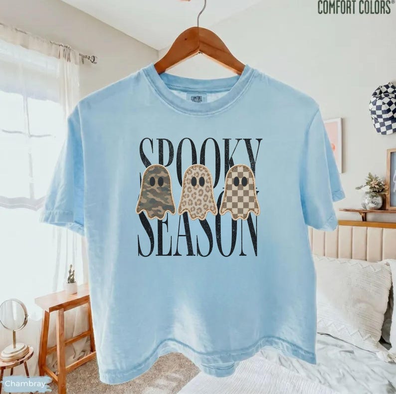 May include: A light blue short-sleeve t-shirt featuring the black text SPOOKY SEASON and three ghost illustrations. The ghosts have distinct patterns: one with camouflage, one with a leopard print, and one with a tan and white checkerboard pattern.