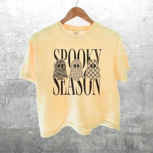 May include: A pale yellow short-sleeve t-shirt featuring the black text SPOOKY SEASON and three ghost illustrations. The ghosts have distinct patterns: one with green camouflage, one with a tan leopard print, and one with a brown checkerboard.
