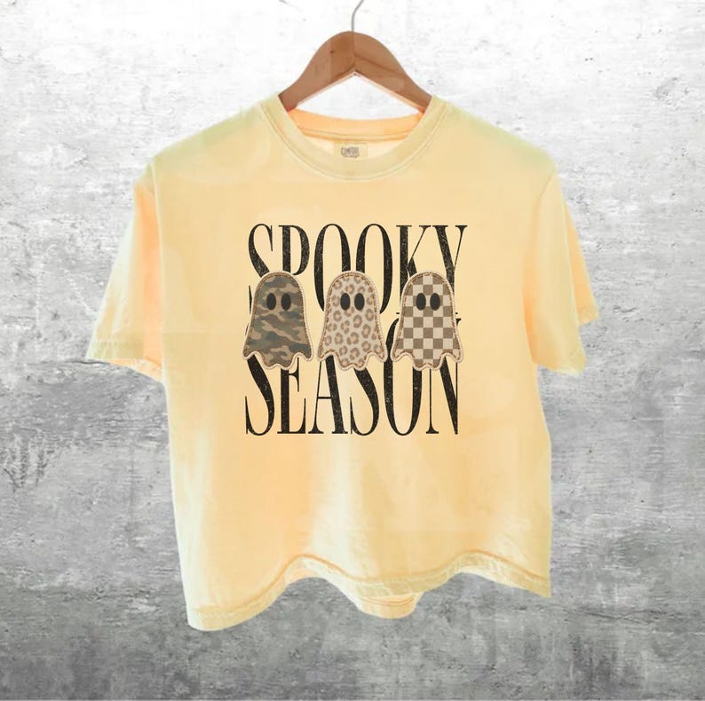 May include: A pale yellow short-sleeve t-shirt featuring the black text SPOOKY SEASON and three ghost illustrations. The ghosts have distinct patterns: one with green camouflage, one with a tan leopard print, and one with a brown checkerboard.