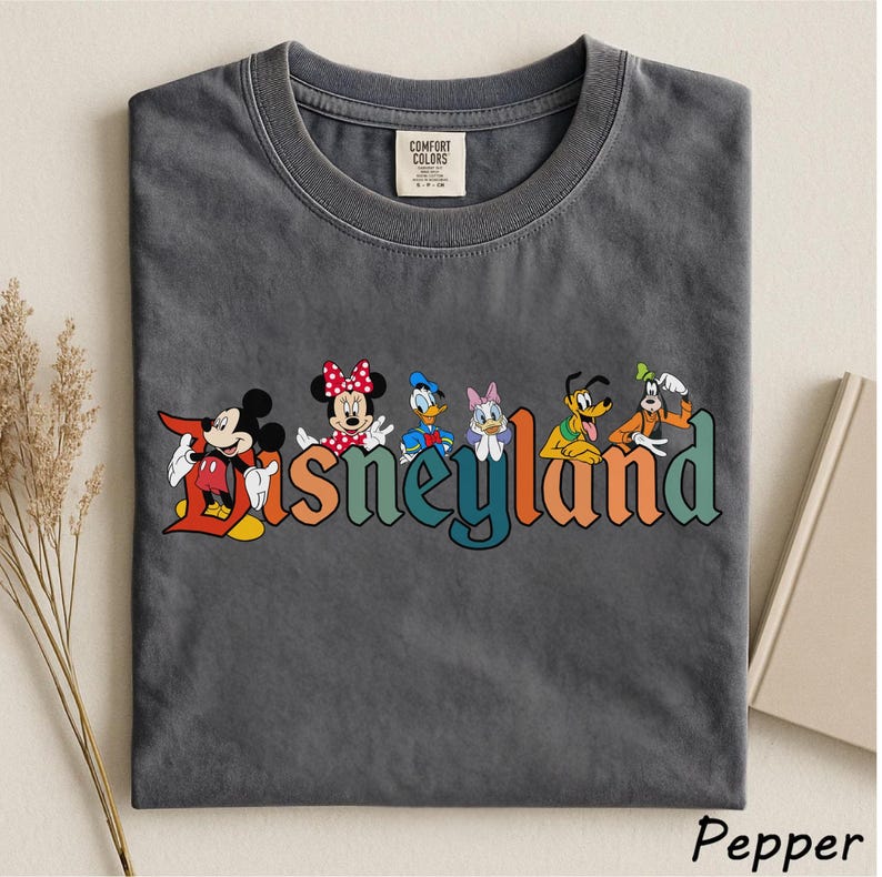May include: A dark gray t-shirt featuring the word "Disneyland" in colorful letters with Mickey Mouse, Minnie Mouse, Donald Duck, Daisy Duck, Pluto, and Goofy. The shirt is made of a soft material and has a classic crew neck.
