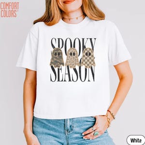 May include: A white short-sleeved t-shirt featuring the black text SPOOKY SEASON. Below the text are three ghost illustrations with different patterns: one in camouflage, one in leopard print, and one in a brown and white checkerboard pattern.
