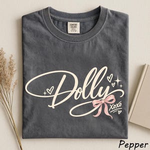 May include: A charcoal gray crew neck t-shirt featuring the word Dolly in a large, cream-colored cursive script. The design includes a small pink bow, several tiny hearts, and star accents, with the text XOXO printed in a smaller font below.
