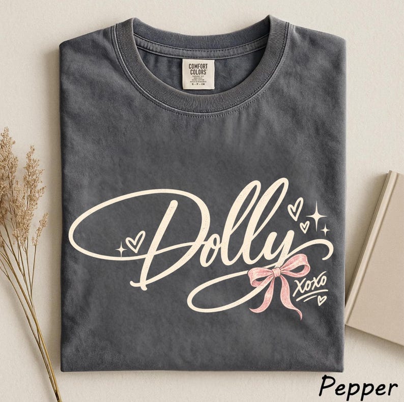 May include: A charcoal gray crew neck t-shirt featuring the word Dolly in a large, cream-colored cursive script. The design includes a small pink bow, several tiny hearts, and star accents, with the text XOXO printed in a smaller font below.